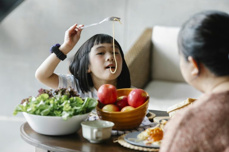 Cute Asian little girl eating delicious spaghetti while sitting at table with fresh fruits and salad with unrecognizable grandmother